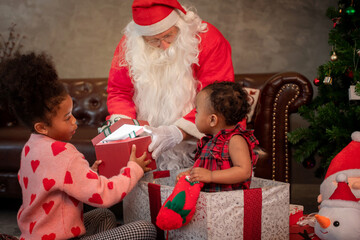 African American sisters in red dresses sit on the floor with Santa Claus at home, paying attention to the gift box in hand, celebrate Christmas