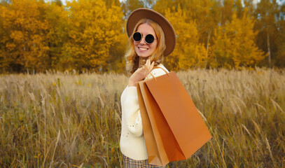 Wall Mural - Stylish happy woman standing with shopping bag in autumn park, fashionable girl smiling outdoors