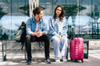 © Iryna - Young Caucasian man and woman sitting on bench at travel station with luggage. Casual outfits and relaxed expressions suggest travel, adventure, and companionship.