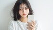 © Sumet - A young woman with dark, wavy hair holds a smartphone, gazing softly at the camera against a minimalistic white background.