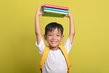 A smiling happy Asian schoolboy holding books on his head isolated on yellow background. Copy space. Happy child Back to School.