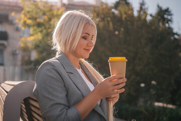 Wall Mural - A blonde woman sits on a bench drinking coffee from a yellow cup. She is wearing a gray jacket and has her hair in a ponytail. The scene is peaceful and relaxing.