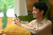 © saltdium - A Young adult man relaxed individual leans against a yellow pillow, smiling while engaged with their smartphone, Engaged in a Moment of Digital Connection