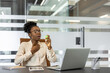 © Liubomir - African American businesswoman at office desk uses throat spray while working on laptop. Health care during workday, with phone and notebook nearby, portraying focus and wellness.