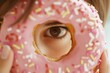 © woters - A child holds a pink frosted donut with sprinkles up to their eye while smiling playfully indoors during the afternoon