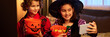 © pressmaster - Panorama of happy girl in Halloween costumes sitting on bed, holding buckets with candies and taking selfie on smartphone