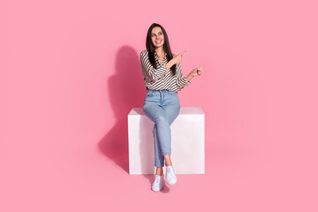 Charming young woman in casual fashion sitting on white cube against pink background, smiling and pointing with both hands