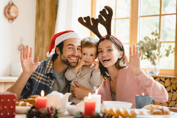 Happy young caucasian family of three parents and toddler celebrating New Year Christmas together at home, spending time on holiday dinner looking at camera