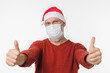 © InsideCreativeHouse - Grey-haired man in red Santa Claus hat wearing protective face mask against coronavirus Covid 19 showing thumbs up isolated in white background. Pandemic Christmas New Year concept