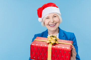 Happy mature woman in Santa hat holding red gift box isolated over blue background. Happy holiday! Happy New Year Eve celebration! Merry Christmas! Winter holidays concept