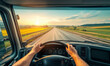 © IBEX.Media - Driver's point of view from inside a truck cabin, hands on the steering wheel, looking out onto a long open highway at sunrise, surrounded by picturesque fields and a clear horizon
