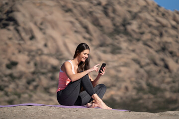 Woman sitting cross-legged on yoga mat, smiling while using her smartphone, enjoying a moment of digital relaxation.