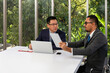 © Meng_Dakara - Two businessmen sitting at table in modern office, one holding tablet, while the other works on laptop, engaged in discussion