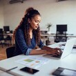 © LIGHTFIELD STUDIOS - A young multiracial businesswoman working at her desk in a contemporary office, with a focus on her reviewing reports and using technology, demonstrating professional dedication