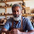 © LIGHTFIELD STUDIOS - A close-up portrait of an older multiracial male in his 60s, sitting at a desk in his small local workshop, surrounded by handmade goods. His thoughtful expression highlights the pride and