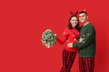 Poster - Happy young couple in pajamas and with Christmas wreath on red background