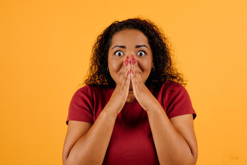 Surprised emotional cute attractive young black woman with bushy hair covering mouth, looking at camera, isolated on yellow studio background. Excitement, shock human emotions concept. High quality