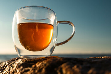 Glass mug of hot beverage resting on rock overlooking water at sunset