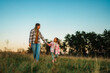 © DusanJelicic - A mother and daughter walking hand in hand through a grassy field at sunset, with trees in the background.