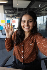 Wall Mural - Latino businesswoman waving at camera during video call. Friendly smile, brown polka dot shirt highlight welcoming appearance. Concept of communication, connection, and professional interaction.