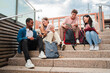 © Jose Calsina - Group of diverse young students talking and smiling while sitting on university campus stairs. Multiracial friends enjoying casual conversation, holding tablets and smartphones in a relaxed academic
