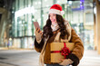 © Home-stock - Modern holiday shopping. Joyful lady in Santa hat holding gift box and using smartphone while standing outdoors of shopping mall, buyer browsing Xmas sales on phone