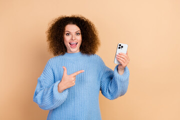 Wall Mural - Photo of blogger curly hair woman wearing blue knitted sweater pointing finger demonstrate smartphone isolated on beige color background