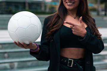 Beautiful brunette businesswoman posing and smiling with joy. Wearing a suit and showing a soccer ball. Leaving work to train and play football. Outdoors. Sports and wellness advertising.