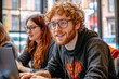 © EJManzaneque - A red-haired man with glasses and a beard works with a laptop in a coworking space with other people. Concept of teamwork in coworkings and startups.