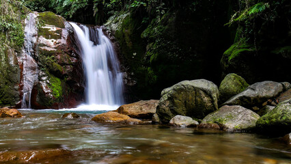 A small waterfall flows rapidly in the middle of a lush tropical forest, surrounded by moss-covered large rocks and green trees in Mount Halimun Salak National Park, Bogor, Indonesia.