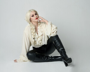 Canvas Print - full length portrait of beautiful female model, wearing vintage gothic fantasy ruffle blouse white shirt, leather pants, blonde hair holding knife weapon. sitting pose, isolated on studio background.