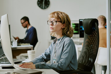 Medium shot of young Caucasian female specialist sitting at workplace in modern office looking through data on desktop computer screen