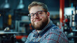 © Nattapol - smiling young man in glasses, wearing checkered shirt and overalls, exudes confidence in workshop filled with tools and equipment. His cheerful demeanor adds warmth to industrial setting