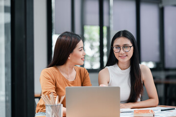 Two young women engaged in a discussion while working on a laptop in a bright, modern office environment.