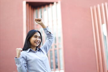 close up of smiling asian businesswoman wear formal suit celebrating winning victory standing over urban red building, female entrepreneur raising fist say yes gesture standing outdoors