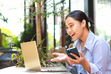 excited asian woman working on laptop and holding smartphone working from outdoor cafe have online meeting with explaining gesture