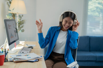 Businesswoman is enjoying music through headphones while working at her desk in a modern office, expressing happiness and taking a break from her tasks