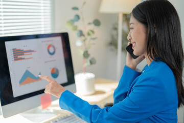 Confident businesswoman in blue suit analyzing financial data on computer screen in office, discussing with phone in hand. Smiling, showing success and professionalism in modern workplace