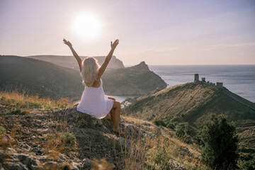 Wall Mural - Happy woman is sitting on a hillside, wearing a white dress. She is surrounded by a beautiful landscape, with a body of sea in the background. Concept of peace and happiness.