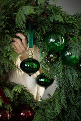 A festive hand delicately hangs a green ornament on a lush evergreen wreath adorned with holiday cheer in a cozy indoor setting