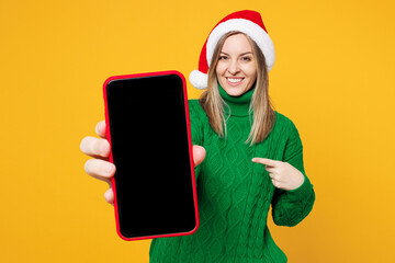 Merry young woman wear green sweater Santa hat posing hold use point finger on blank screen mobile cell phone isolated on plain yellow background. Happy New Year celebration Christmas holiday concept