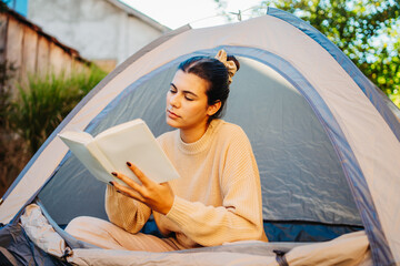 young caucasian woman reading a book in her tent in backyard