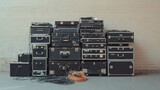 A collection of black equipment cases with cords and wires spilling out in front of a white brick wall.