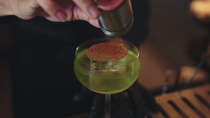 Cropped bartender garnishing caramelized citrus slice on cube ice in green Midori Sour cocktail served in stylish stemmed glass. Perfect for sophisticated bar settings christmas tree theme. Vinosity.