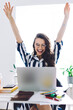 © BullRun - Young happy female worker sitting at desk with laptop and holding hands up