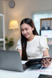 ©  NCST Studio - Young asian businesswoman holding a document and smiling while working on a laptop in her home office