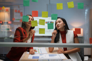 Two businesswomen are having a brainstorming session in the office at night, using sticky notes on a glass wall