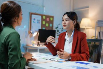 Two businesswomen having a discussion about work while sitting together at a desk in a modern office late at night