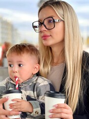 A mother and a child are drinking a drink in a cafe. A young woman in glasses drinks coffee, a baby sits on her arms and drinks milk through a tube.