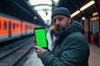 © PinkiePie - Man holding a phone with green screen while standing at a train station